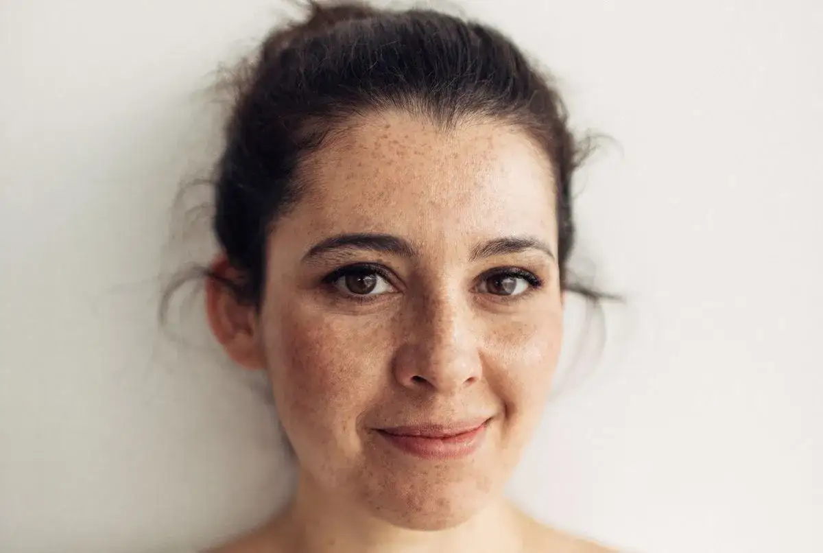 Visage de femme aux taches de rousseur, souriant doucement. Sa peau, bien que présentant une légère **peau terne**, rayonne d'une beauté naturelle.