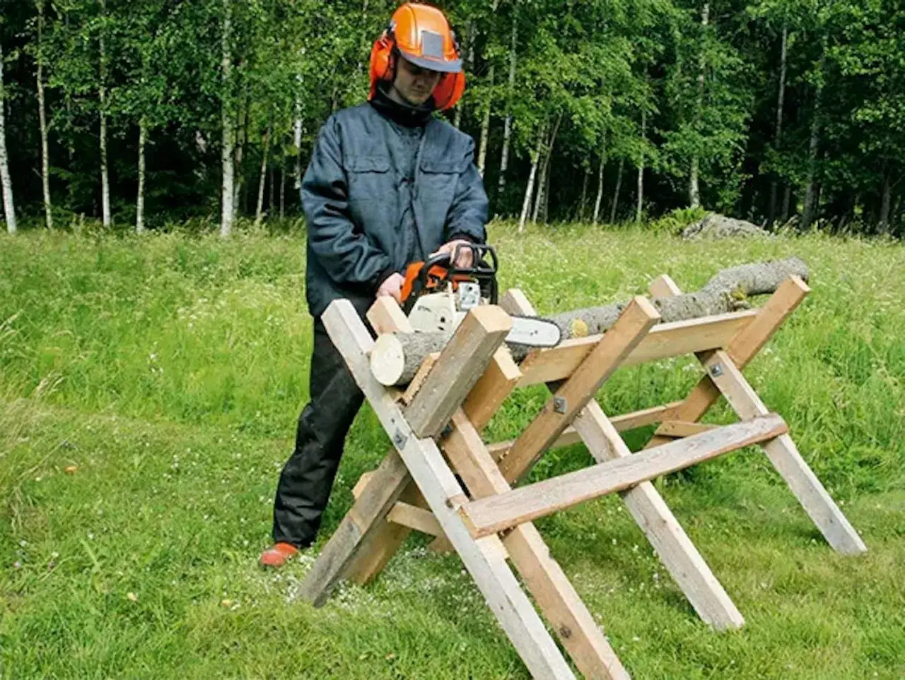 Man i skyddsutrustning använder motorsåg för att kapa ved på en hemmabyggd sågbock i en grönskande skogsmiljö.