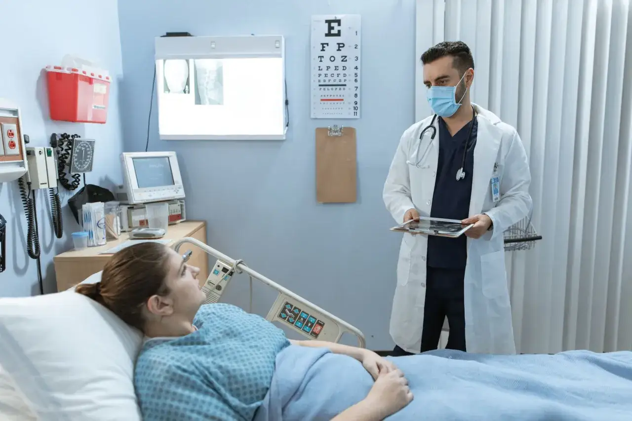 Lekarz w masce rozmawia z pacjentką leżącą w łóżku szpitalnym. Może omawiać zastrzyki na odchudzanie.