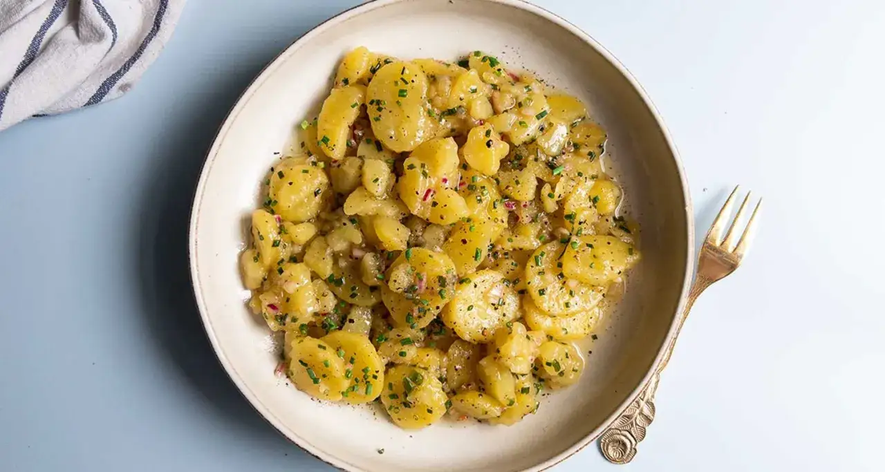Ein köstlicher Kartoffelsalat ohne Mayo, mit frischen Kräutern und Zwiebeln verfeinert, serviert in einer Schüssel mit einer goldenen Gabel.