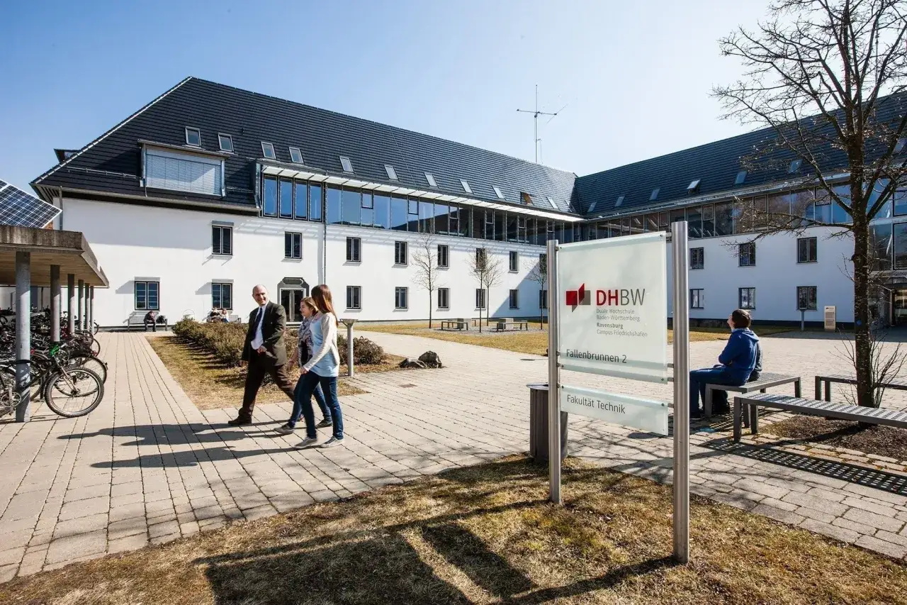 Sonniger Tag am Campus der Hochschule Friedrichshafen. Menschen gehen über den gepflasterten Weg vor dem modernen Gebäude.
