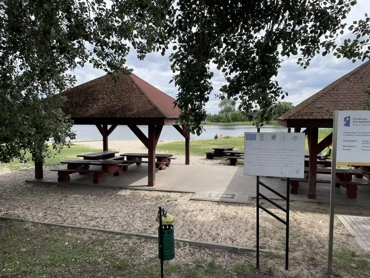 Plaża w Rogoźno Wielkopolskie z wiatami i ławkami. W tle jezioro i wypoczywający ludzie.