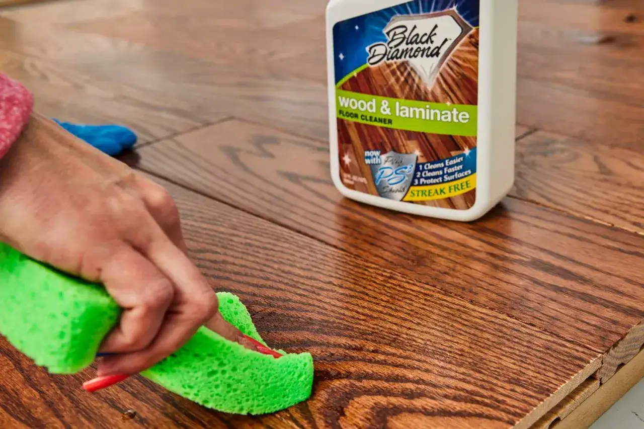 A hand uses a green sponge to clean a wood-look floor, demonstrating how to clean vinyl plank flooring with a Black Diamond cleaner.