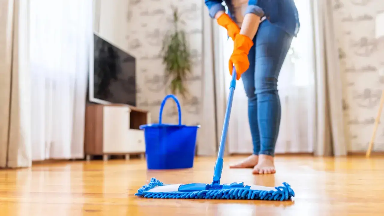 Persona con guantes naranjas y jeans limpia el parquet con una mopa azul. Cubo azul al lado.