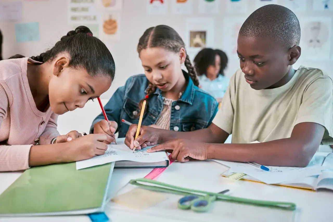 Drei Kinder arbeiten konzentriert an einem Tisch, nutzen kreative Methoden für Gruppenarbeit und schreiben in ihre Hefte.