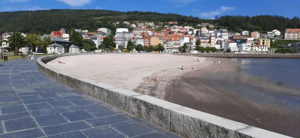 Paseo marítimo y arena dorada en la playa de Cee, con el mar azul y casas blancas al fondo.