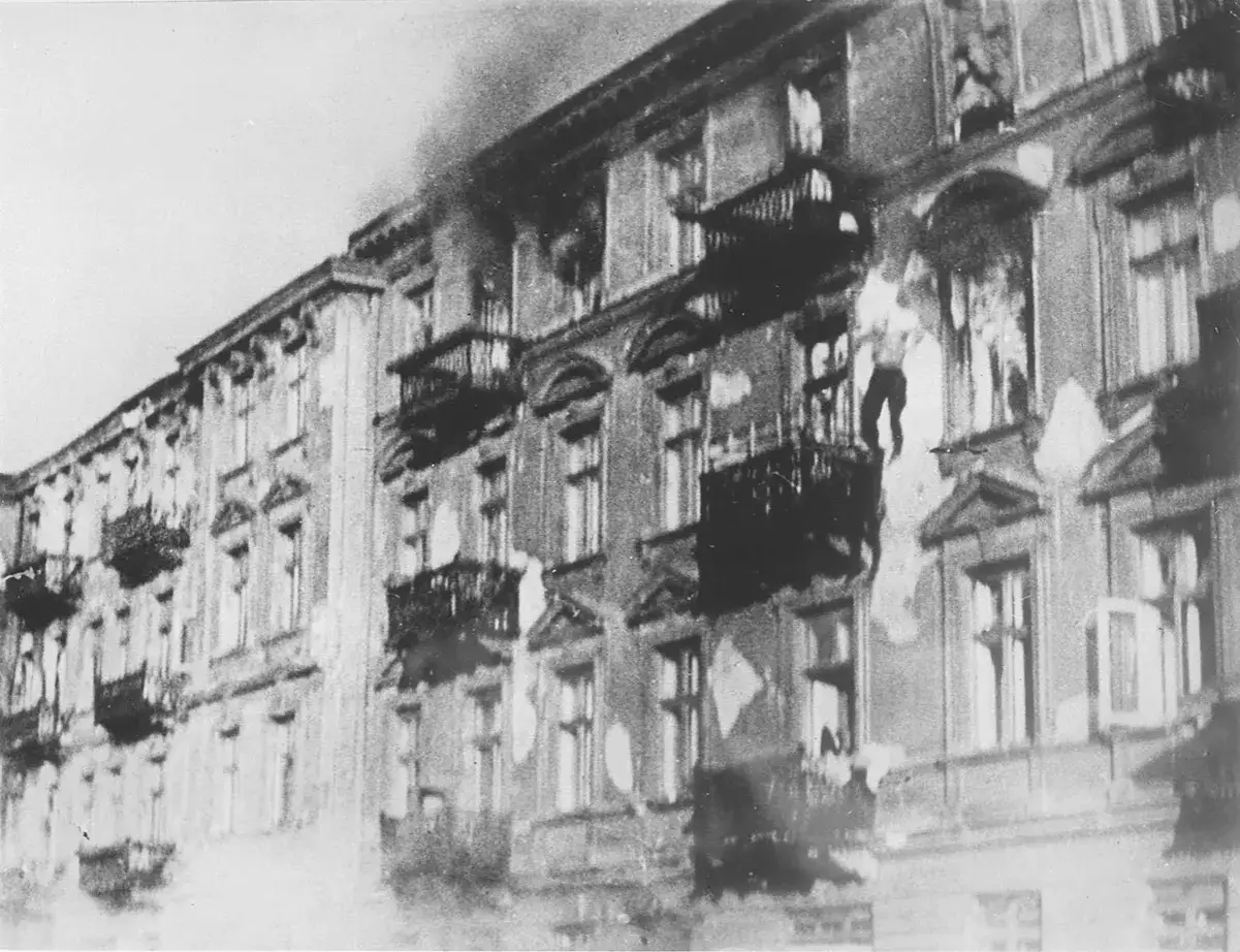 Brennende Geb&auml;ude im Warschauer Ghetto. Rauch steigt auf, Fenster sind zerbrochen, Balkone schwarz. Ein Mann st&uuml;rzt aus einem Fenster.