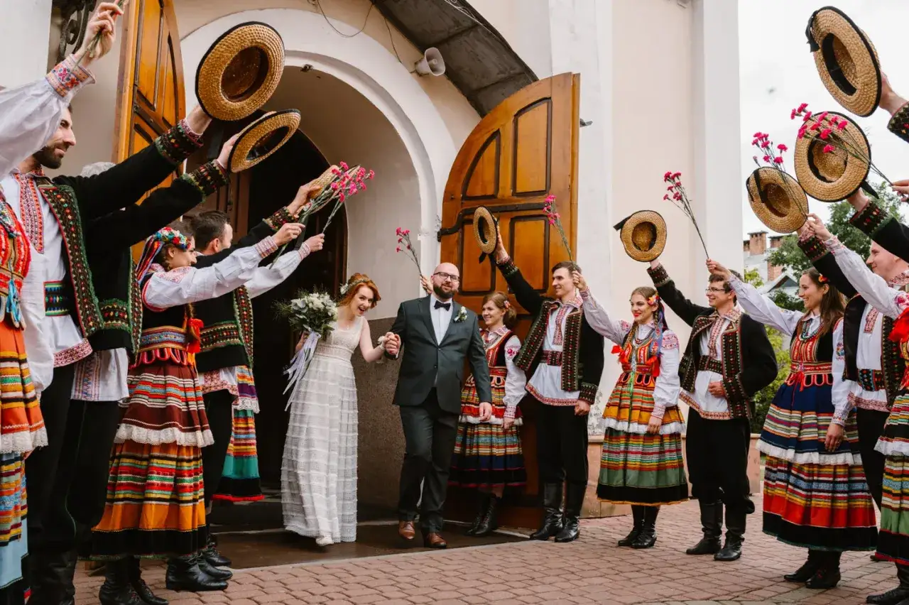 Młoda para wychodzi z kościoła, a goście w tradycyjnych strojach ludowych witają ich, unosząc słomiane **maszki na weselu** i kwiaty.