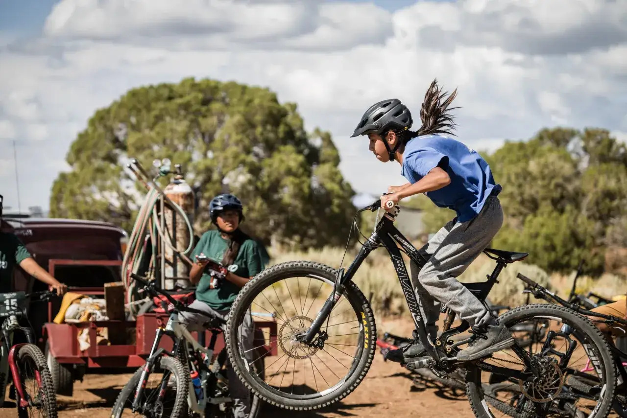 A young girl rides a mountain bike, her hair flying as she navigates a dirt path. Other bikes and people are visible in the background.