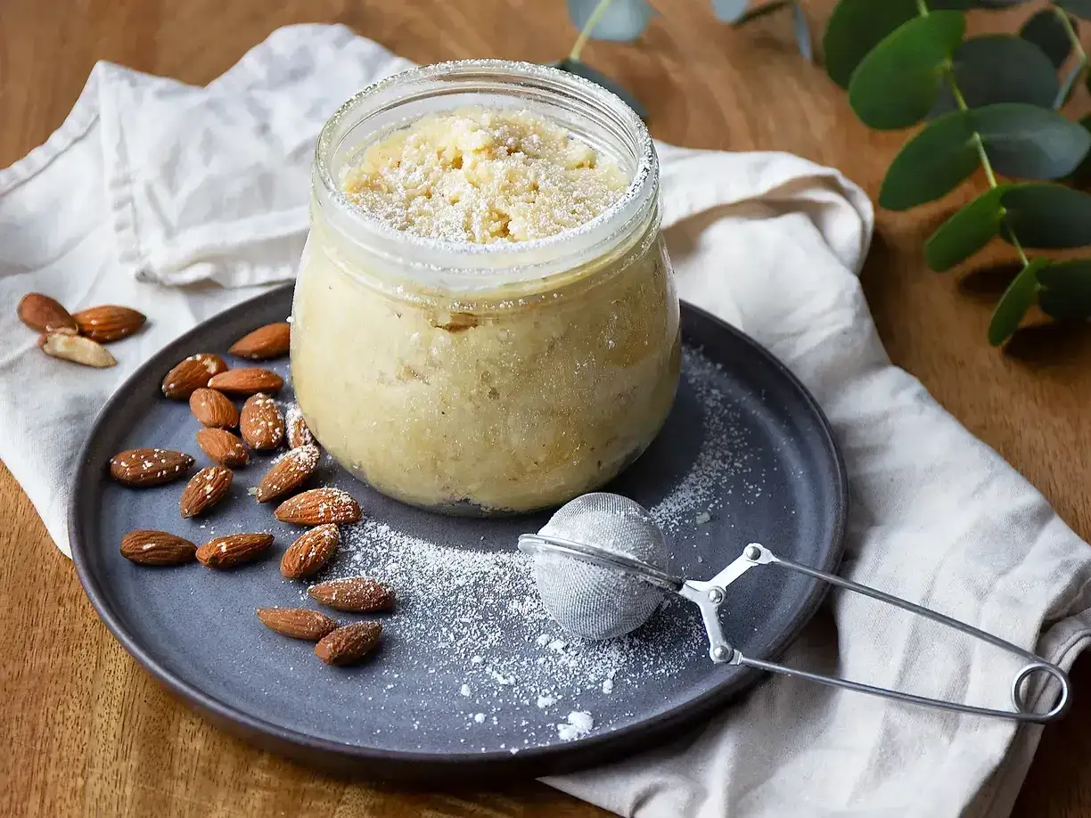En lyxig mandelmassa i burk, toppad med florsocker. Mandlar och en tesil ligger på en tallrik bredvid. Ett perfekt recept för hemmabruk.