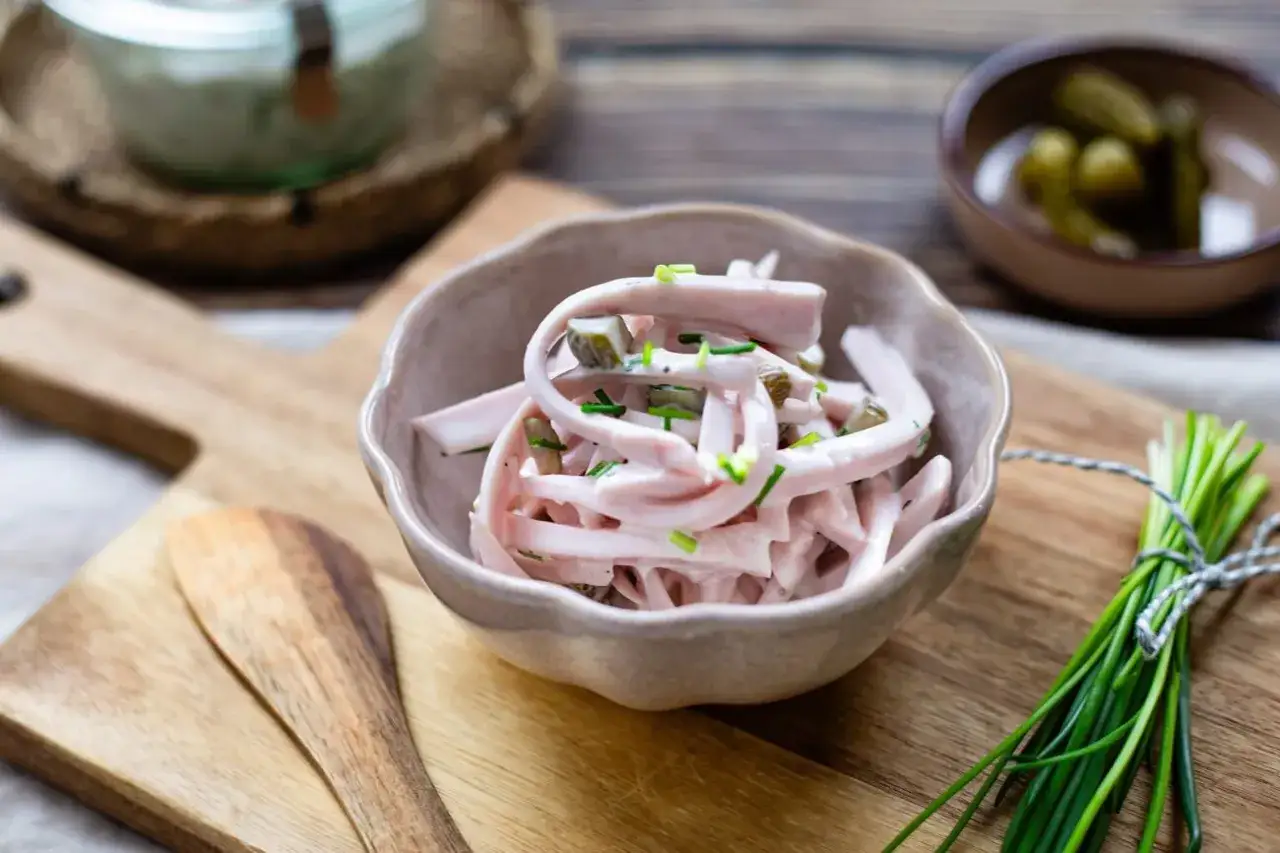 Schmackhaftes Fleischsalat Rezept in einer Schale, garniert mit Schnittlauch. Daneben ein Bund frischer Kr&auml;uter und Gew&uuml;rzgurken.