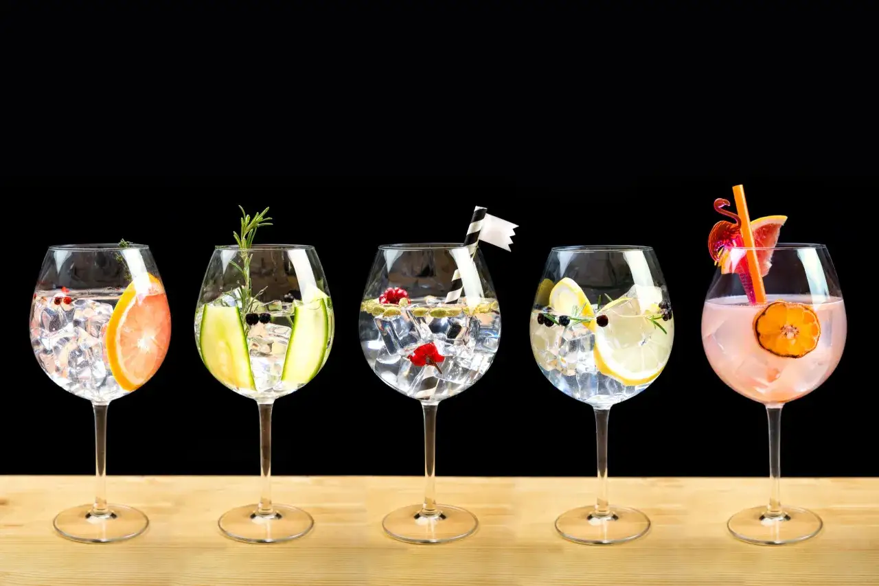Pięć drinków z tonikiem, każdy z innym owocem i ziołami, na drewnianym blacie.