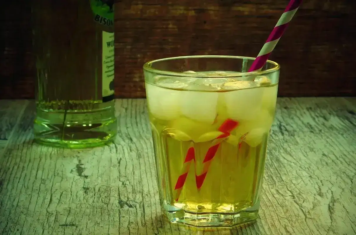 Szarlotka drink przepis: orzeźwiający napój z lodem i słomką, obok butelka z zielonym płynem.
