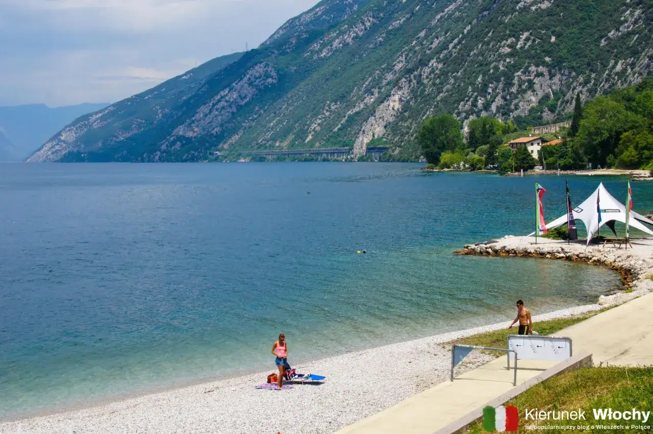 Piękna plaża nad Jeziorem Garda, gdzie **temperatura wody** zachęca do kąpieli. W tle góry i biały namiot.