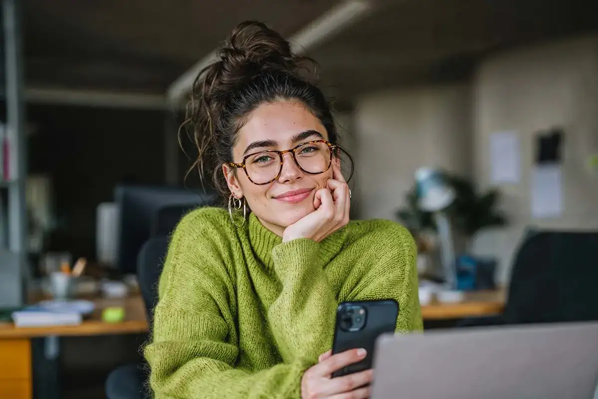 Junge Frau mit Brille und Dutt l&auml;chelt in die Kamera, h&auml;lt ein Smartphone. Sie arbeitet im B&uuml;ro an ihrer IU Kommunikation und PR.