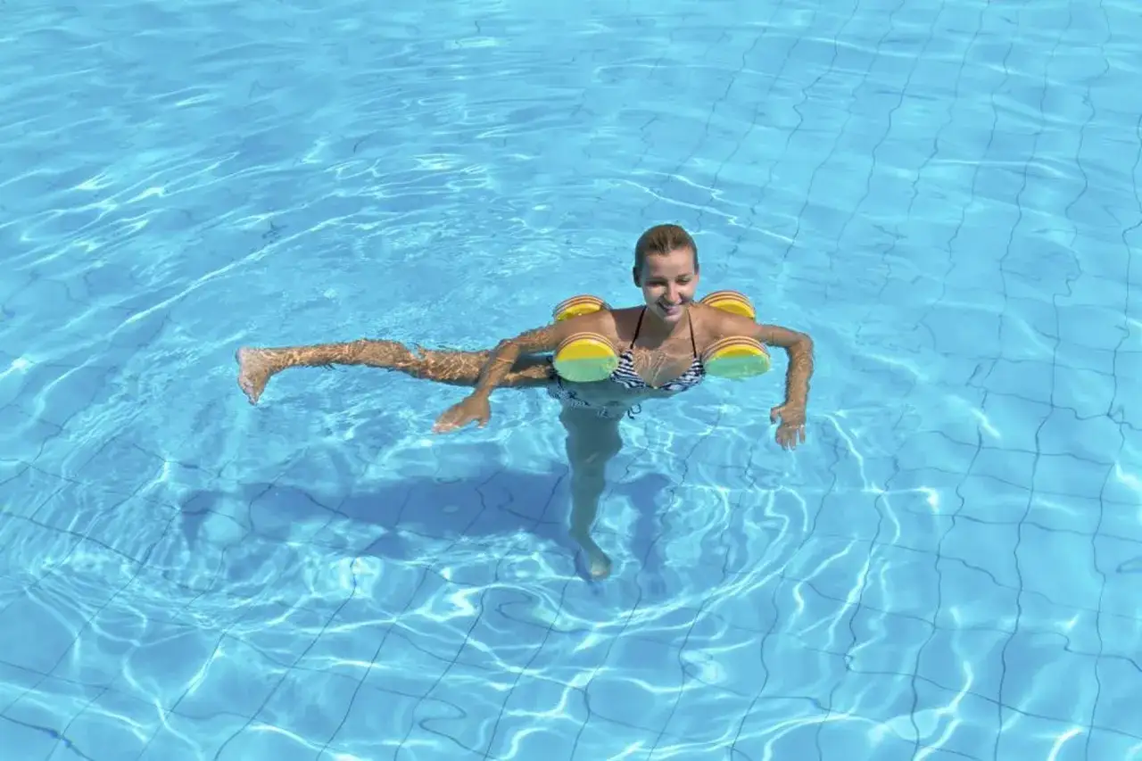 Uśmiechnięta kobieta w bikini wykonuje ćwiczenia w wodzie, unosząc się dzięki żółto-zielonym pływakom.