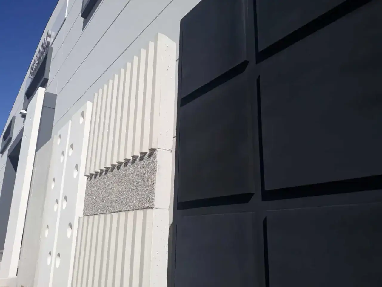 Detalle de una pared prefabricada exterior con texturas y colores contrastantes: paneles blancos acanalados y con c&iacute;rculos, un bloque de hormig&oacute;n granulado y paneles negros cuadrados.