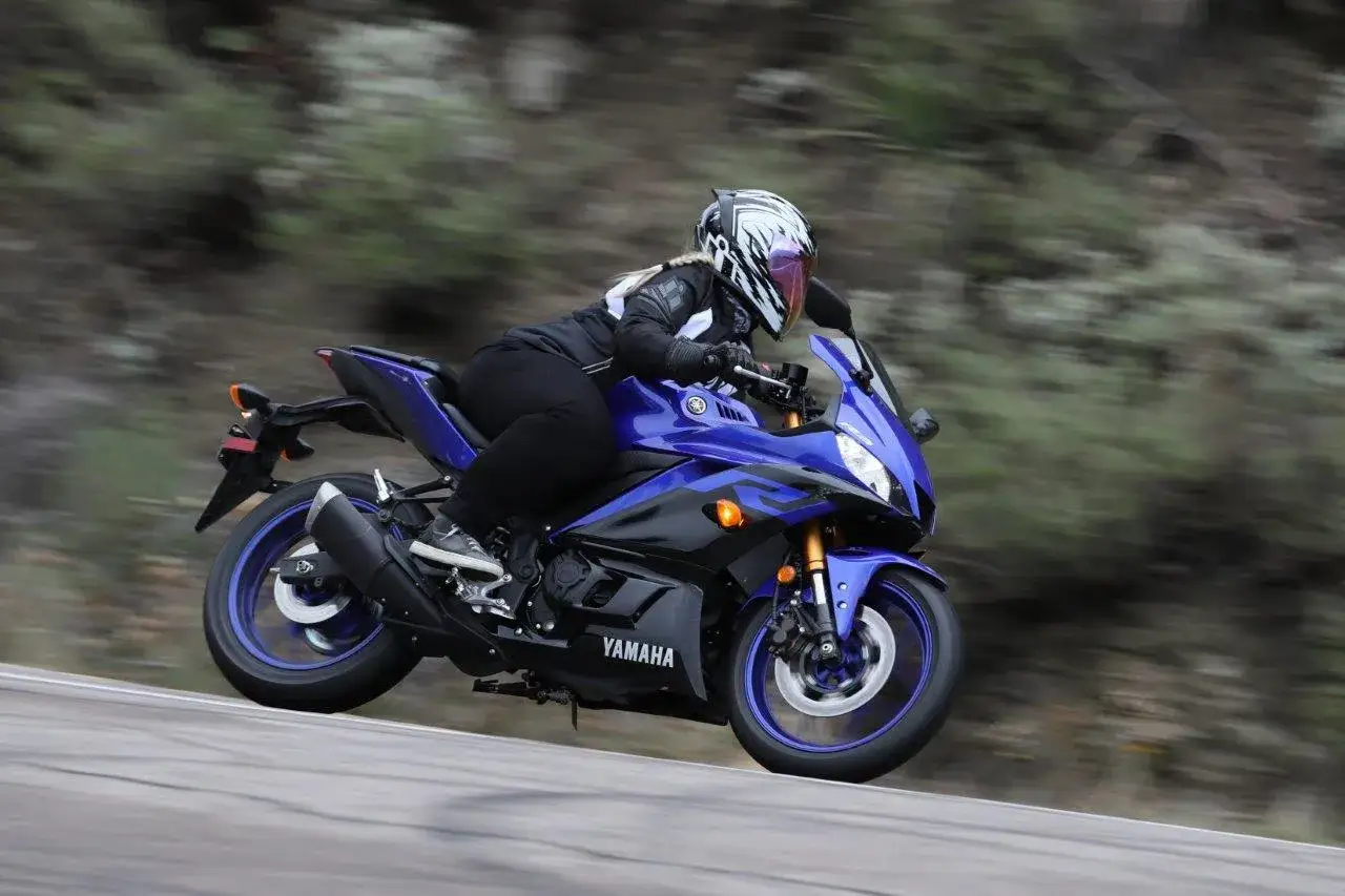 Kobieta w kasku pędzi na niebieskim motocyklu Yamaha r3 po krętej drodze.