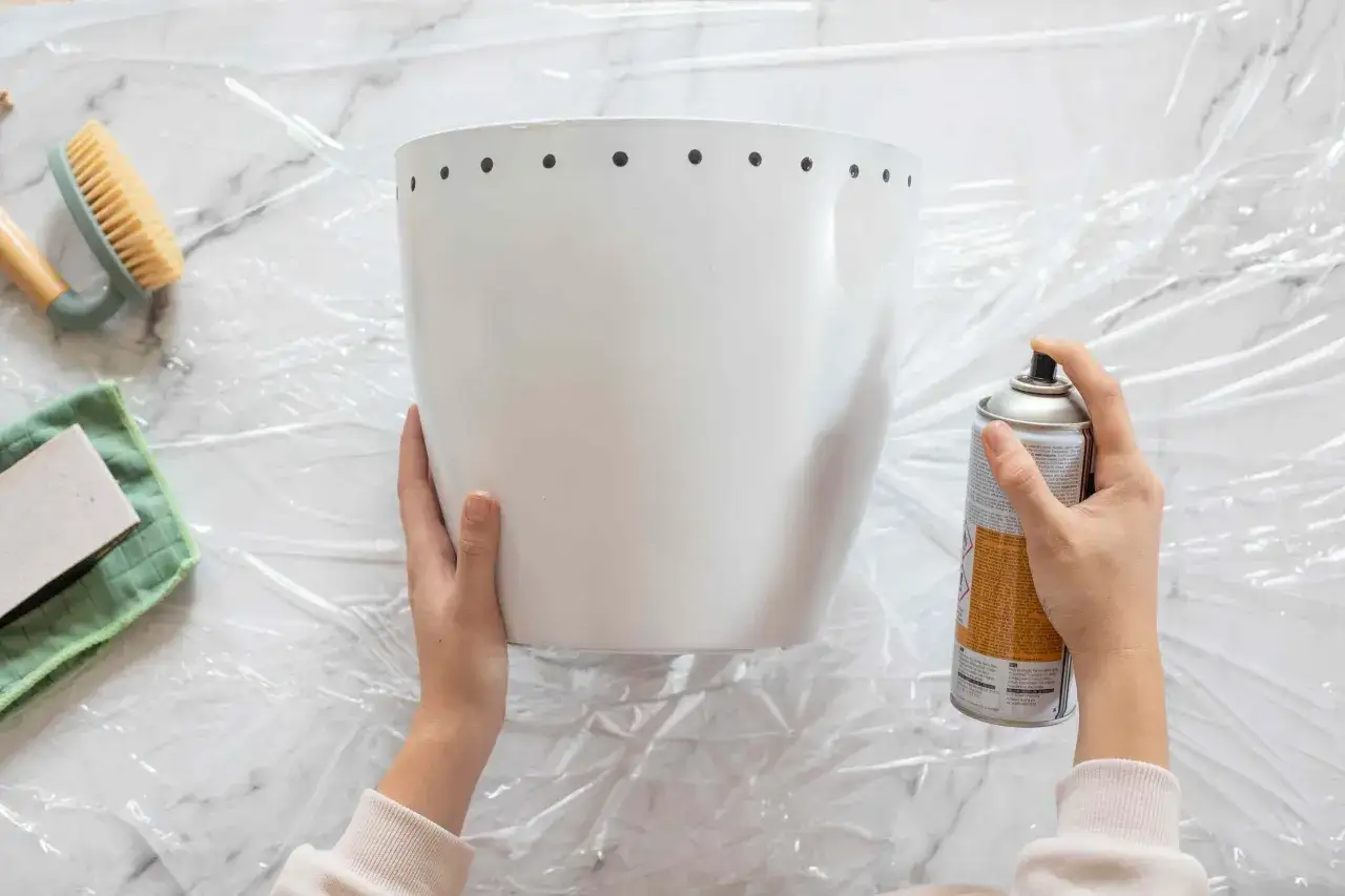 Manos rociando pintura en spray sobre una maceta blanca, prepar&aacute;ndola para un proyecto de como pintar plastico.