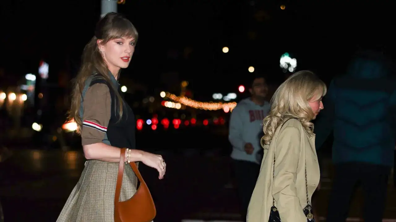 Taylor Swift wzrost: artystka w kratowanej spódnicy i brązowym sweterku, z rudą torebką, idzie ulicą nocą.