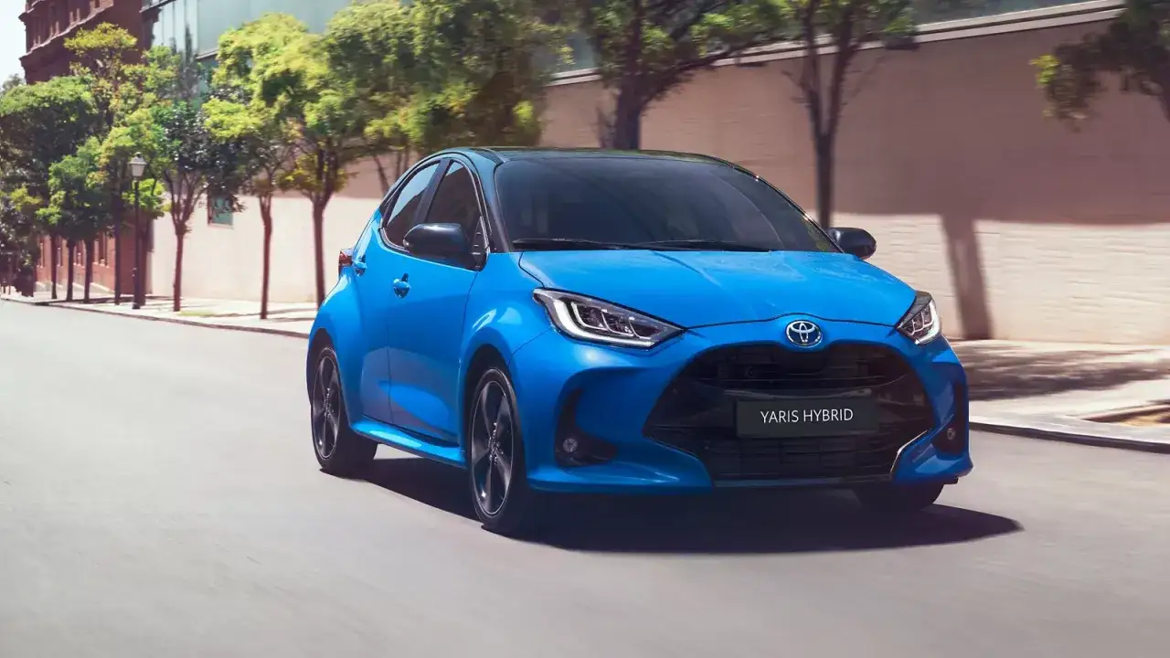 Niebieski Toyota Yaris Hybrid pędzi ulicą, otoczony zielenią drzew i miejską architekturą.