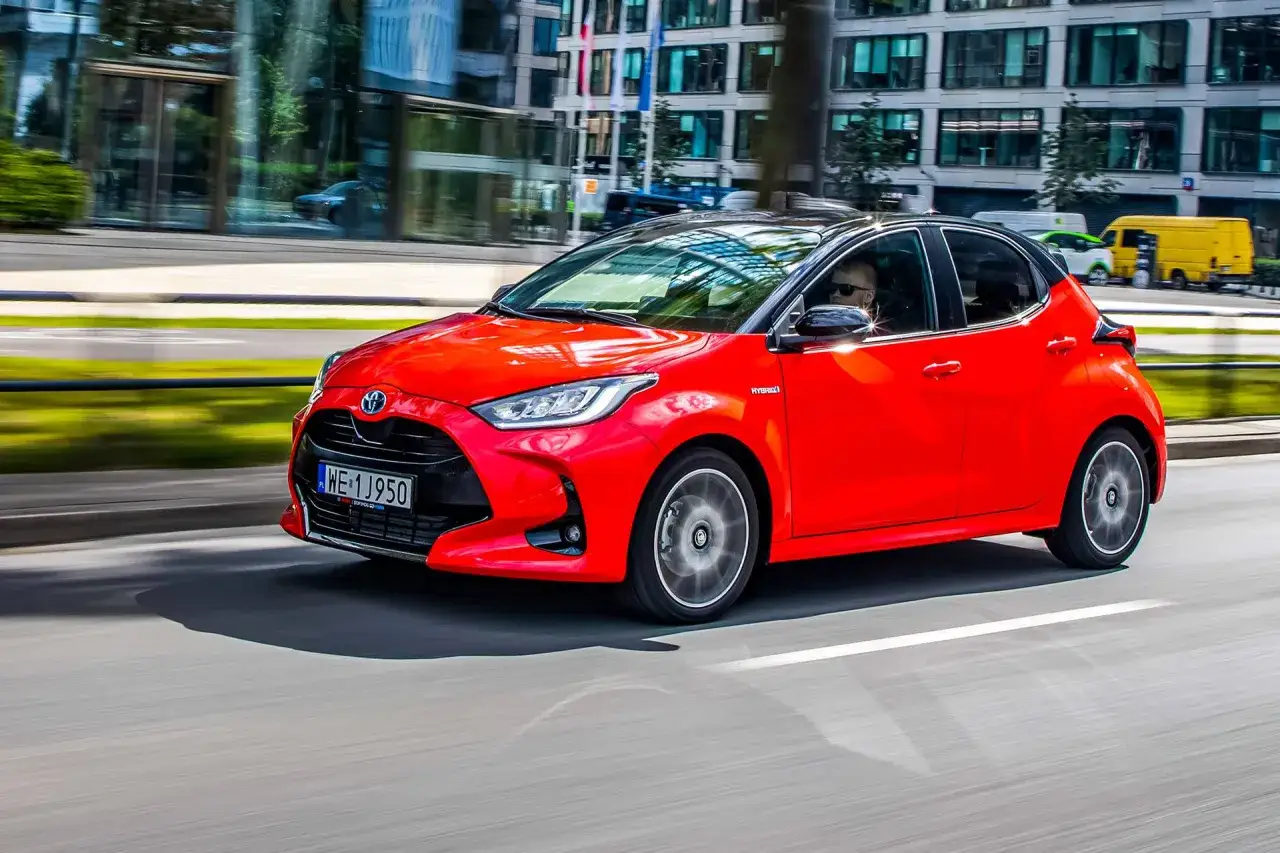 Czerwony Toyota Yaris Hybrid pędzi ulicą miasta. Kierowca w okularach przeciwsłonecznych prowadzi auto.