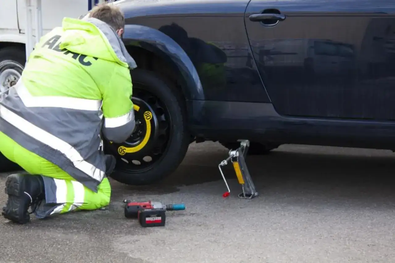Un uomo in giacca catarifrangente cambia una ruota sgonfia dell'auto con l'aiuto di un cric e un avvitatore.