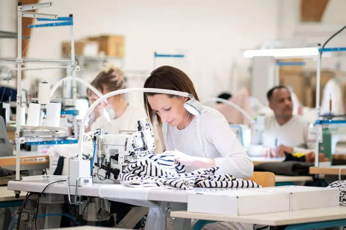 Une ing&eacute;nieure textile travaille avec pr&eacute;cision sur une machine &agrave; coudre, entour&eacute;e de ses coll&egrave;gues dans un atelier lumineux.