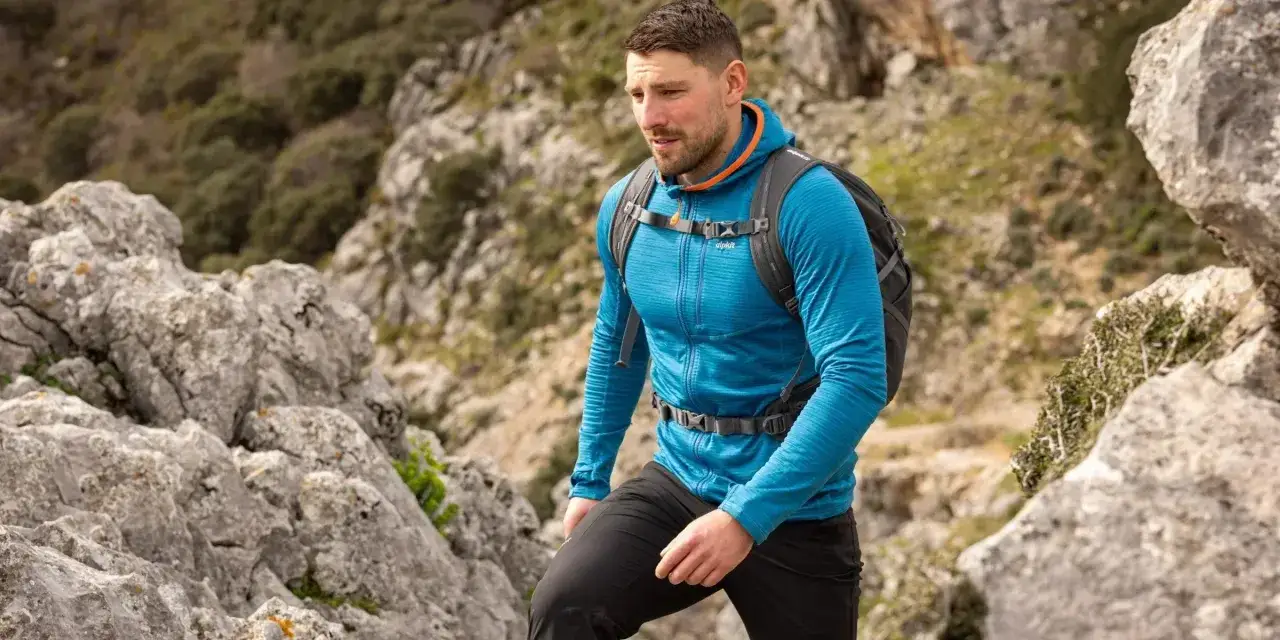 Ein Mann wandert in einem blauen Midlayer mit Reißverschluss und Kapuze durch felsiges Gelände.