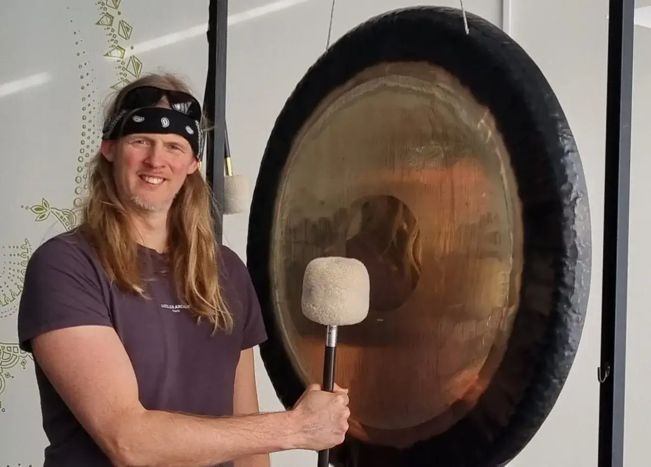 Man med l&aring;ngt h&aring;r och bandana h&aring;ller en gong-klubba framf&ouml;r en stor gong. Hans leende antyder att han &auml;r mitt i en session f&ouml;r gongbad forskning.