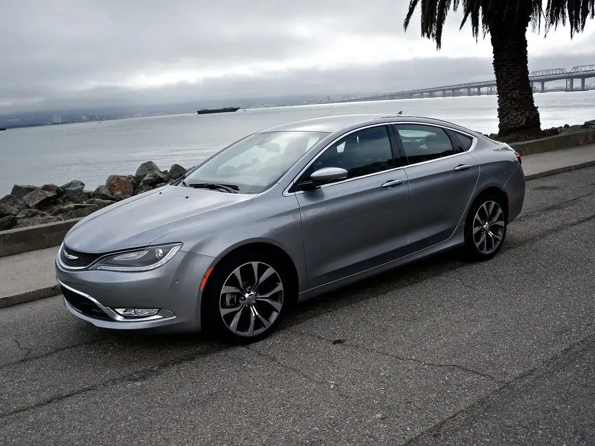 Srebrny Chrysler 200s zaparkowany przy nabrzeżu, z palmą i mostem w tle.