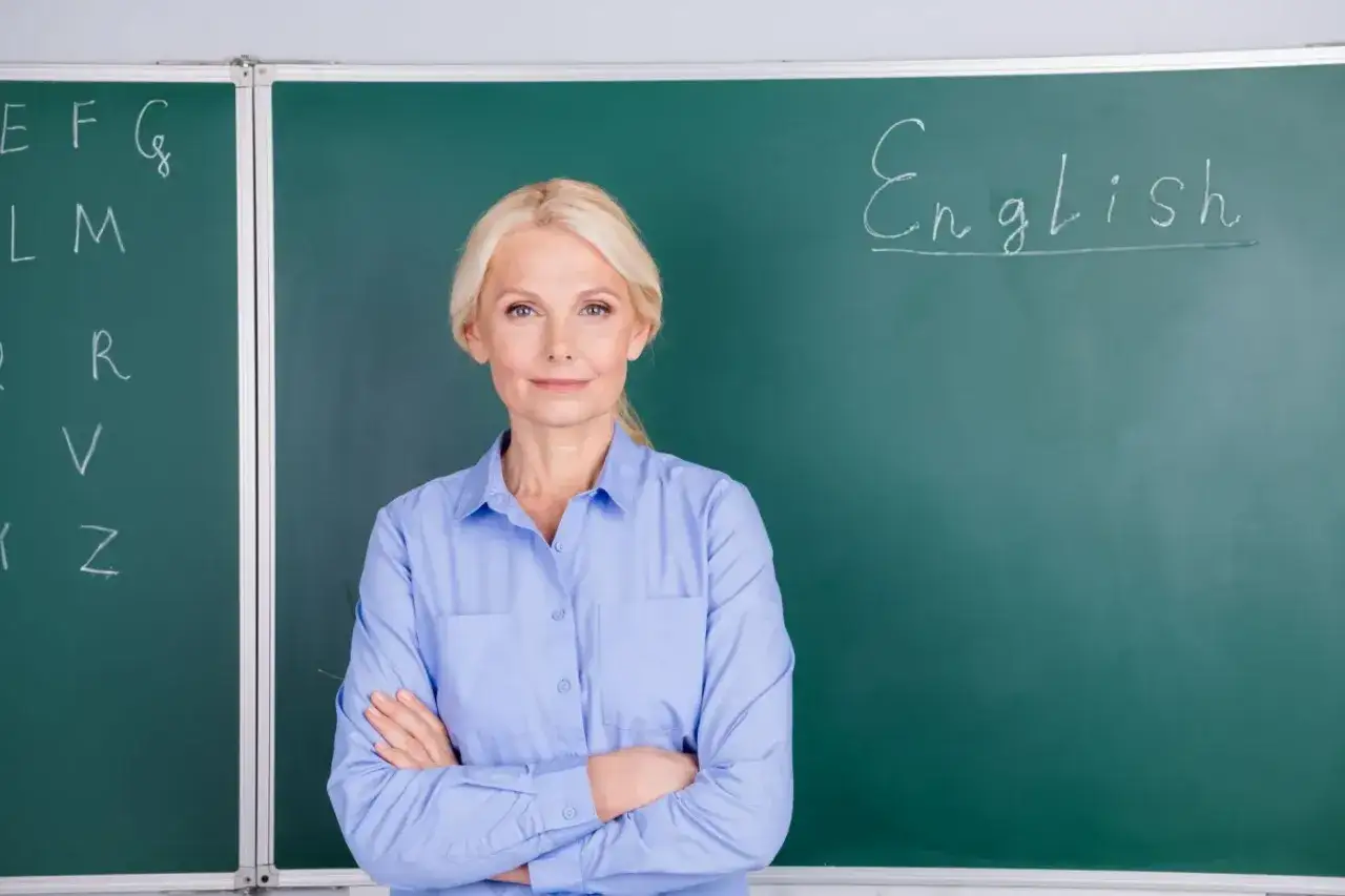 Nauczycielka angielskiego z uśmiechem, marząca o wcześniejszej emeryturze dla nauczycieli. Na tablicy litery i napis