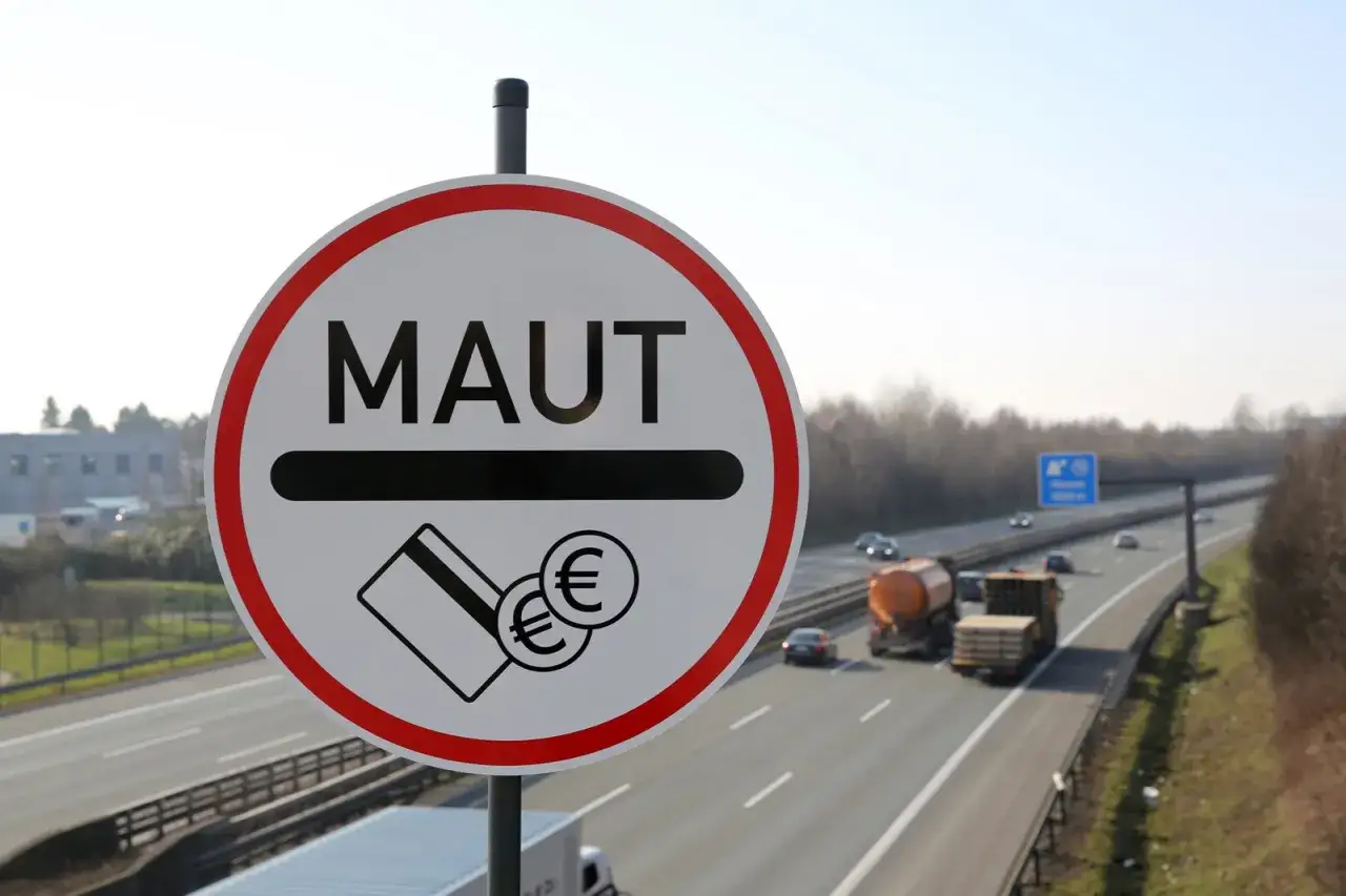 Znak informuje o opłatach drogowych (winieta Niemcy). Na autostradzie widać ruch samochodowy.