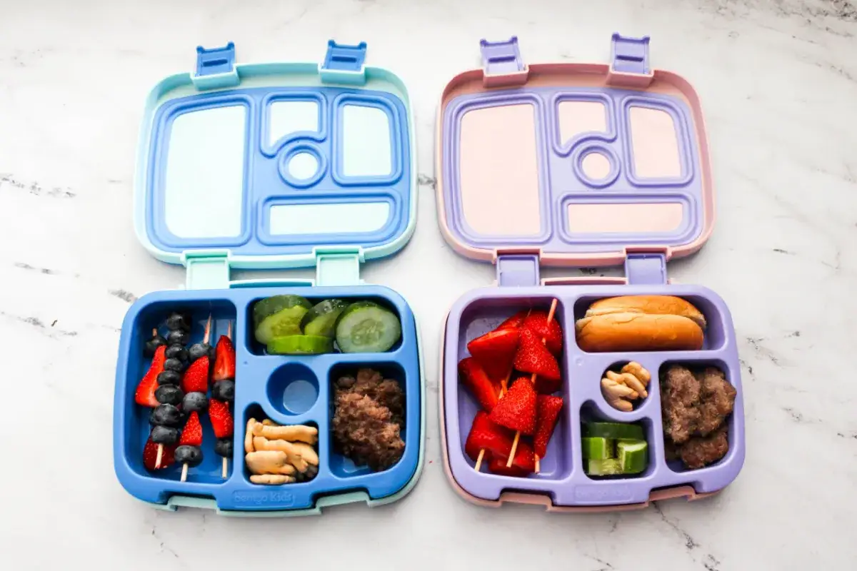Two bento boxes, one blue and one purple, are filled with lunch items. The blue box contains fruit skewers, cucumber slices, crackers, and ground meat. The purple box holds strawberry skewers, a bun, nuts, and cucumber slices.