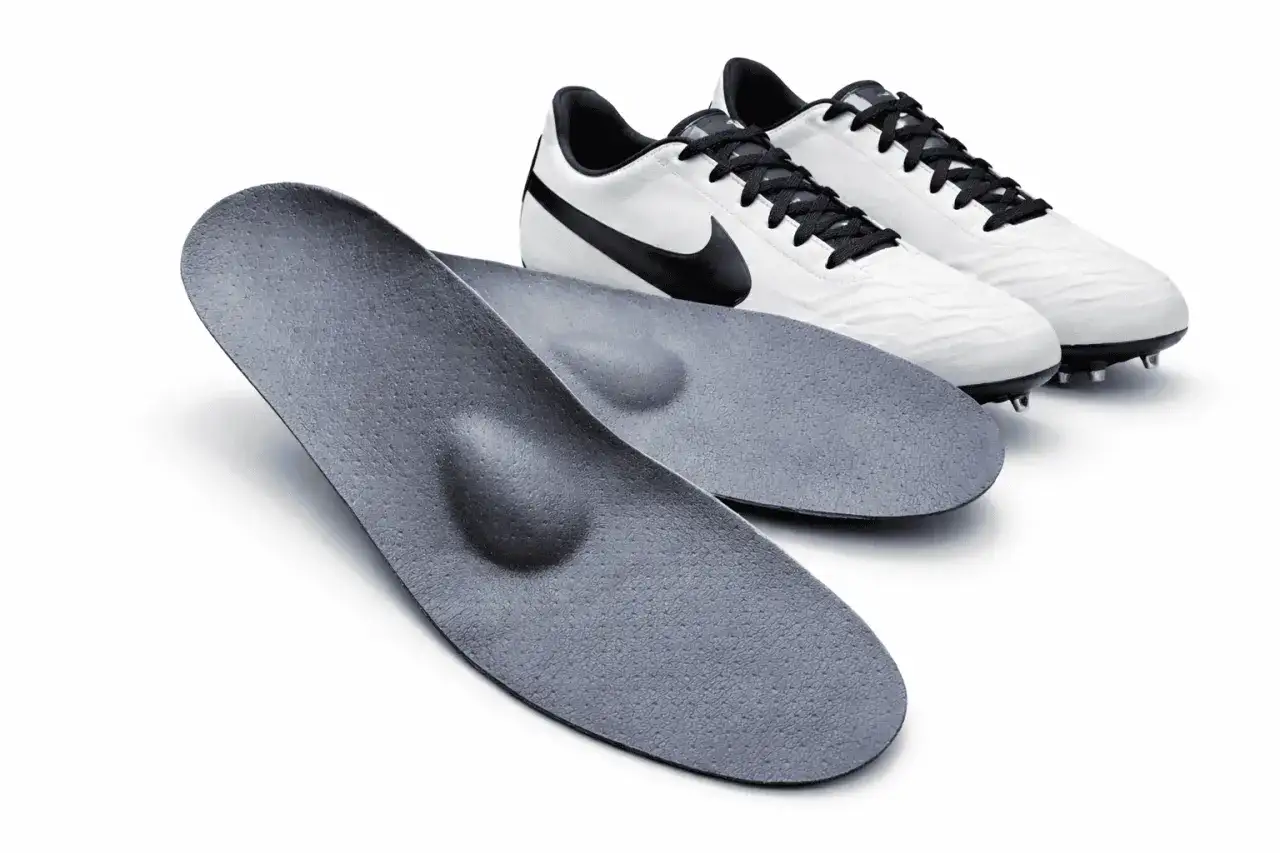Zwei anatomisch geformte Fu&szlig;ball Einlagen liegen vor einem Paar wei&szlig;er Fu&szlig;ballschuhe mit schwarzen Schn&uuml;rsenkeln und Swoosh-Logo.
