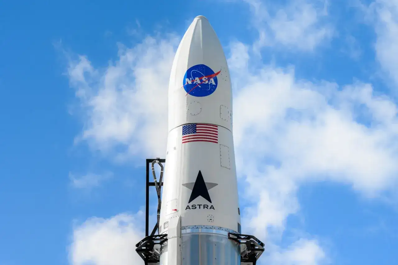 Biała rakieta Astra z logo NASA i flagą USA, gotowa do startu na tle błękitnego nieba z chmurami.