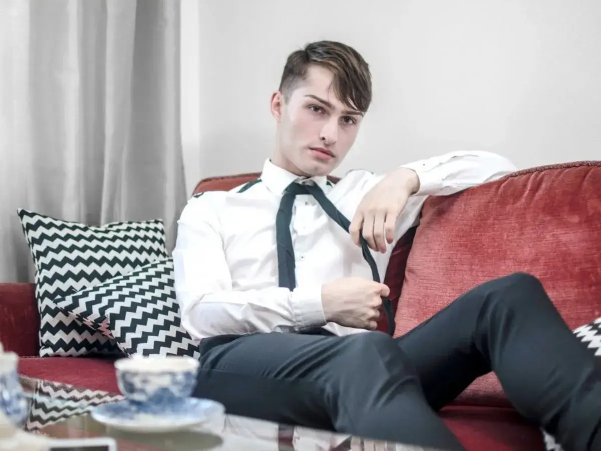 Junger Mann in typisch franz&ouml;sischer Kleidung, wei&szlig;es Hemd, schwarze Hose, sitzt auf rotem Sofa.