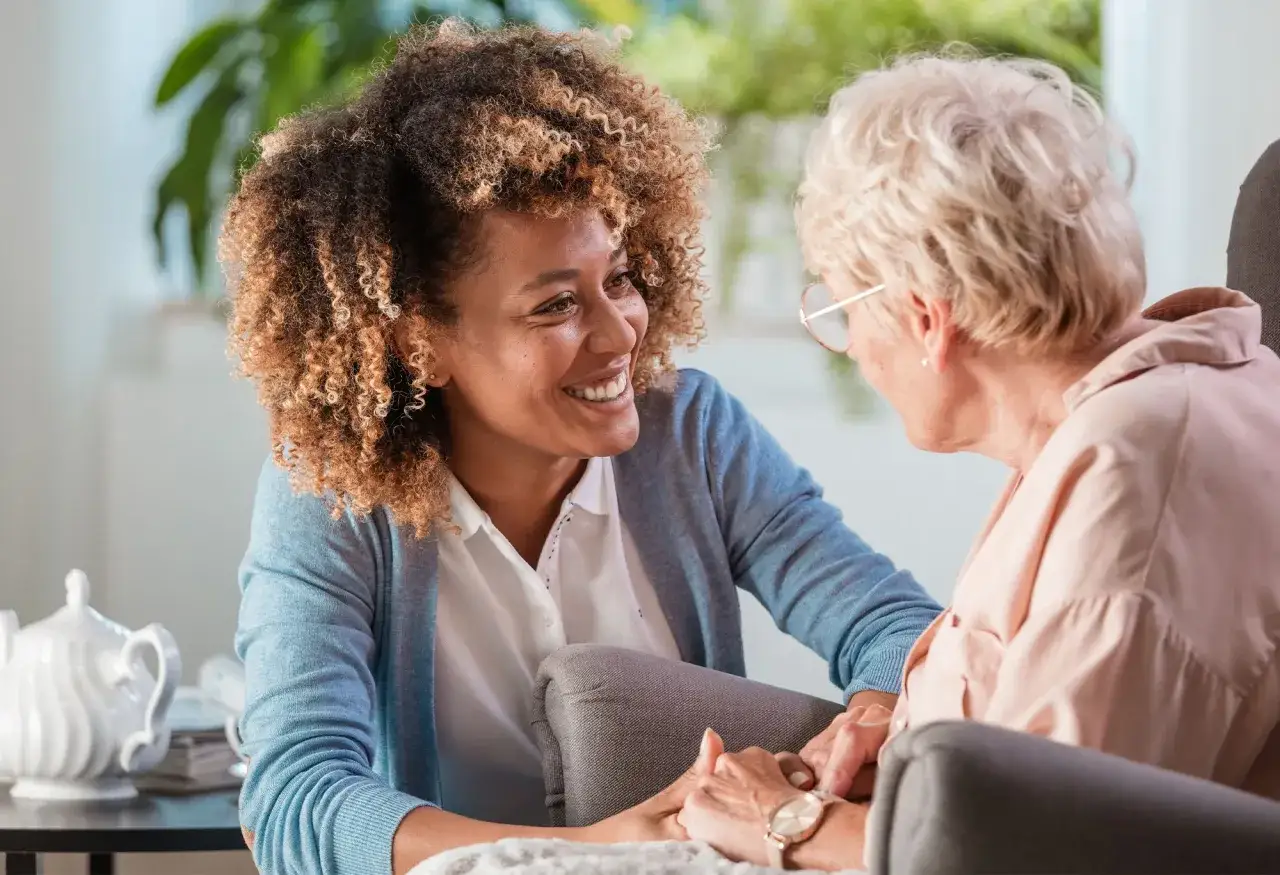Une aide souriante discute avec une personne âgée, main dans la main. Un moment de complicité qui illustre un plan d'aide personnalisé pour personne âgée.