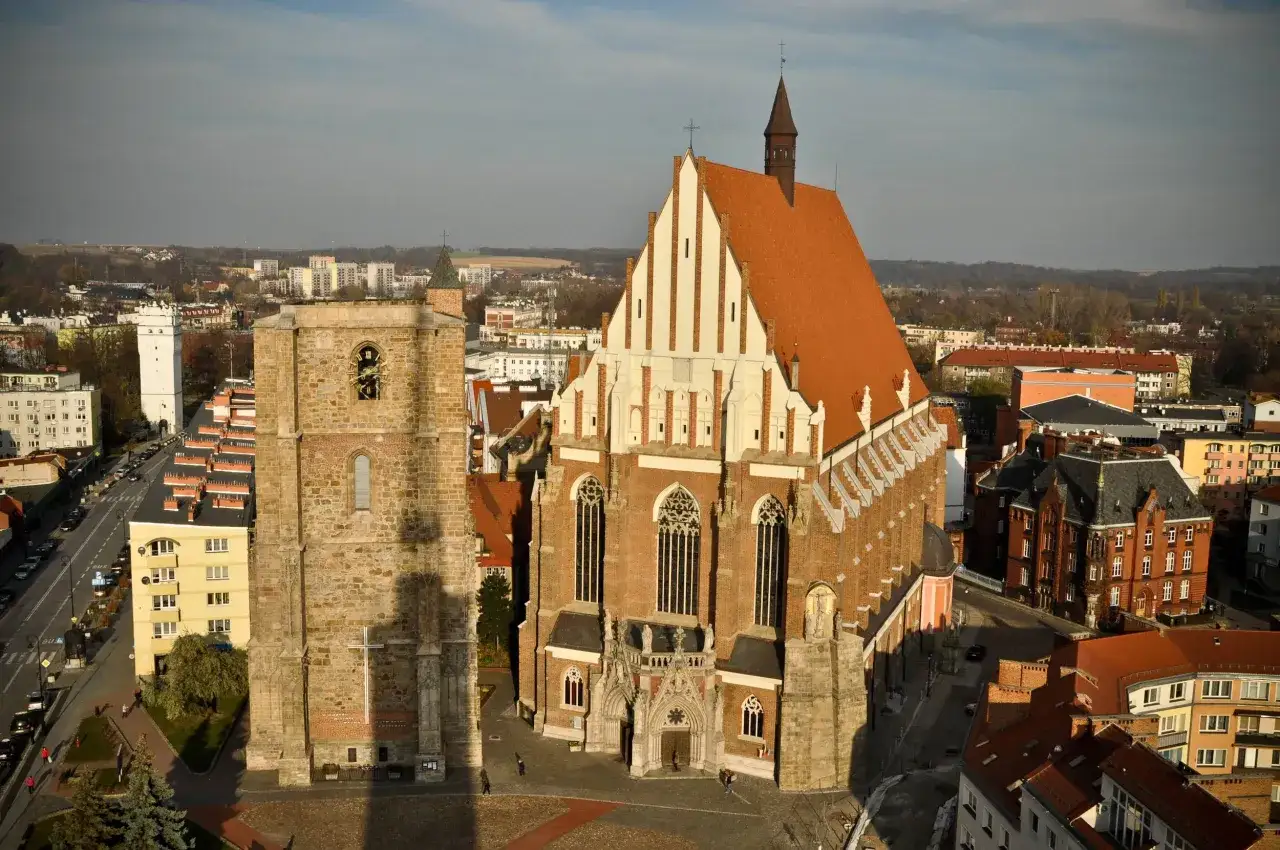 Widok z g&oacute;ry na zabytkowe budowle Nysy. Na pierwszym planie kamienna wieża i ceglany kości&oacute;ł z czerwoną dach&oacute;wką. W tle miejska panorama.
