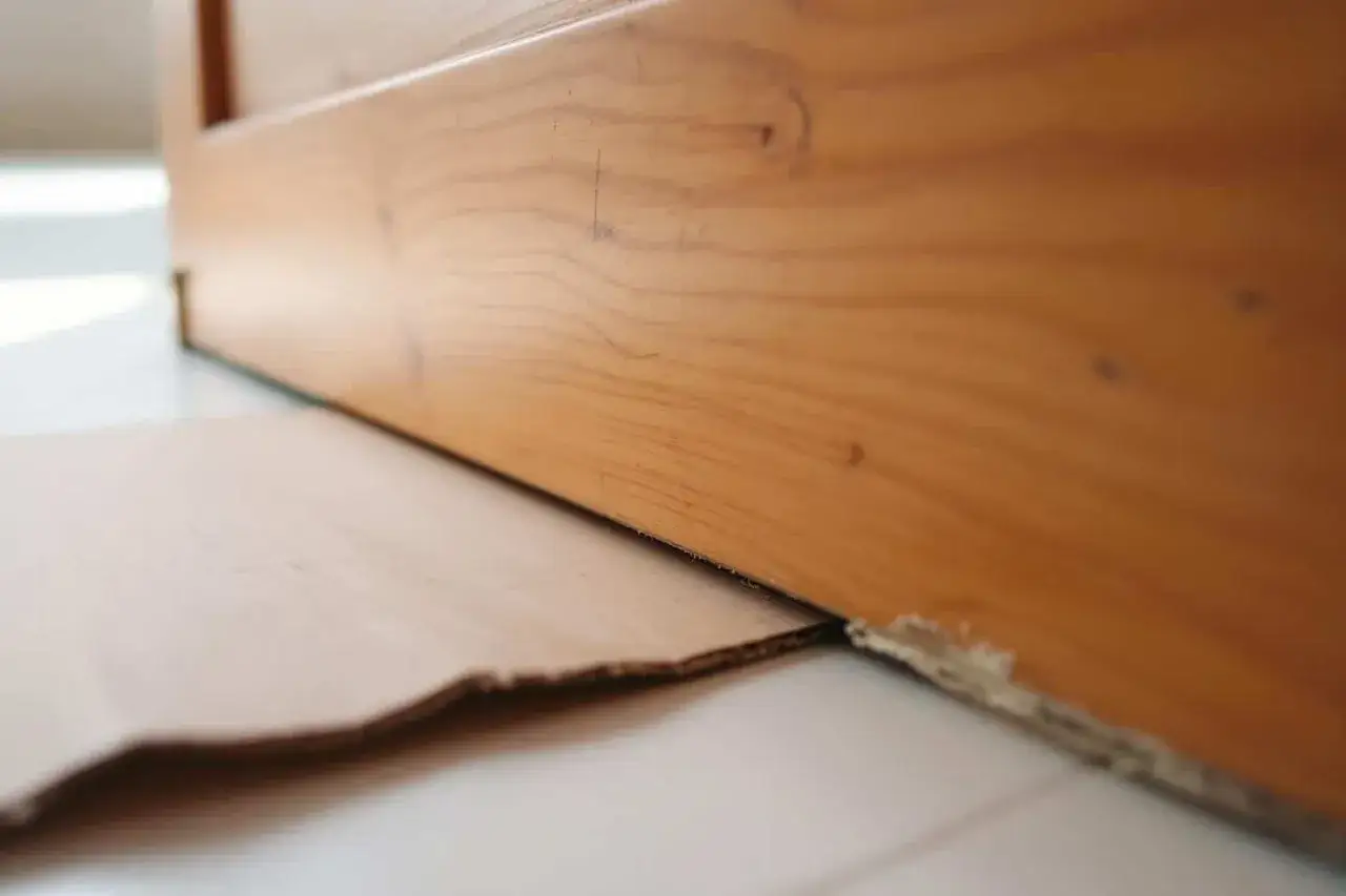 Le bas d'une porte en bois est surélevé par du carton. Le détalonnage porte est visible, avec des marques sur le bois.