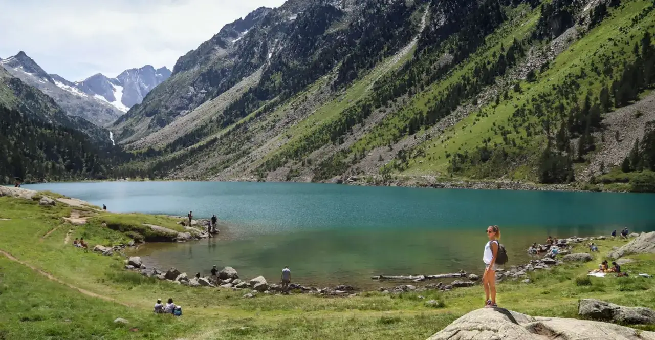 Une jeune femme admire le **Lac de Gaube**, entouré de montagnes verdoyantes et de sommets enneigés. Des promeneurs profitent du paysage.