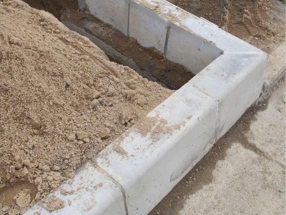 Piaskowy fundament pod szklarnię, wykonany z betonowych elementów i piasku.