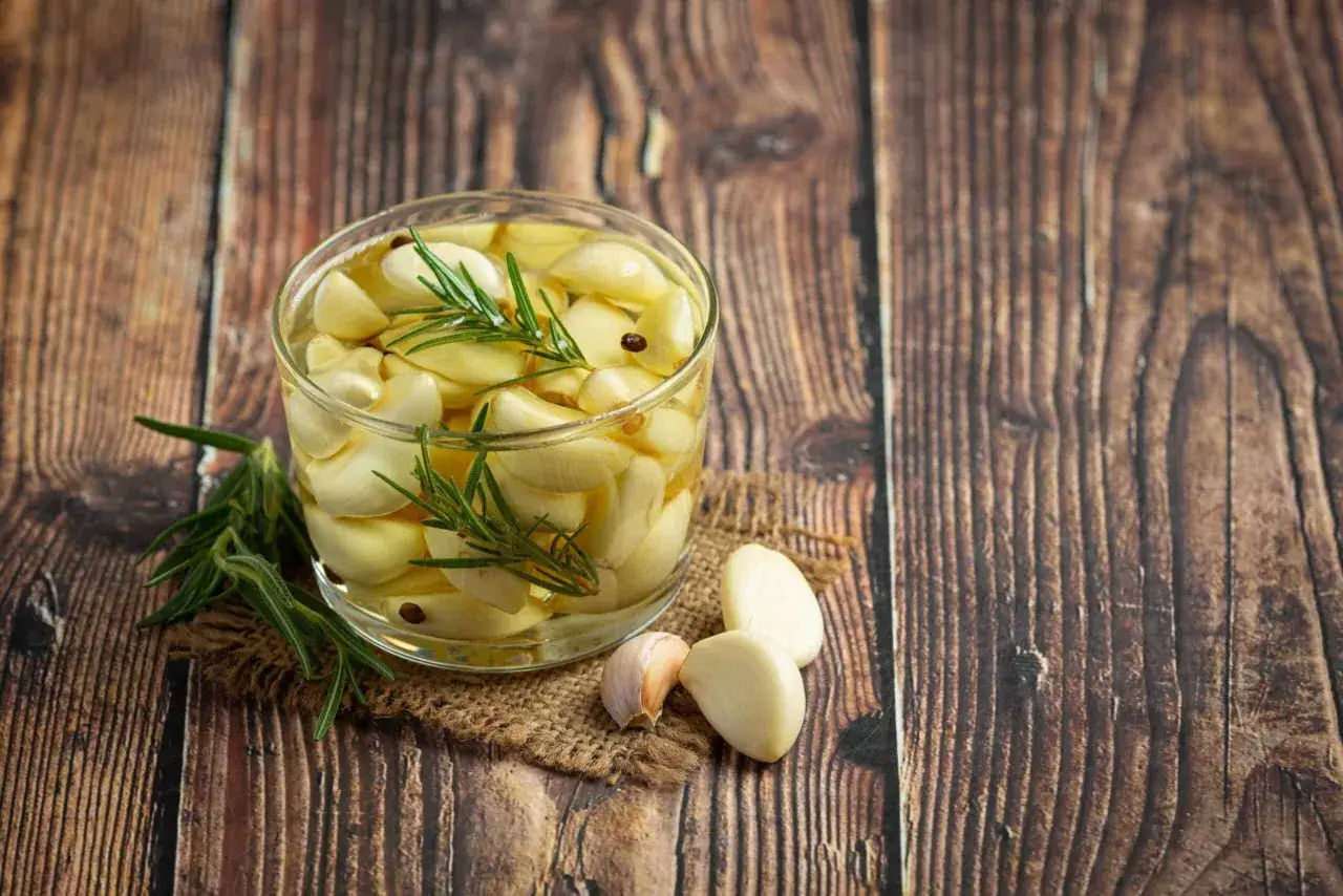 Un bocal de gousses d'ail confit avec du romarin et des grains de poivre sur une table en bois. Une idée pour faire de l'ail confit.