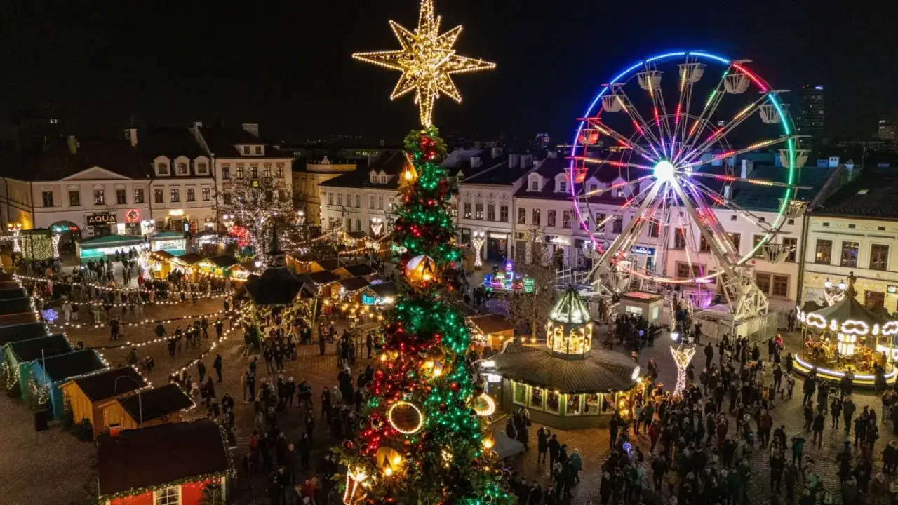 Tłum ludzi spaceruje po placu, podziwiając najpiękniejsze jarmarki bożonarodzeniowe. W centrum stoi ogromna choinka, a w tle widać kolorową karuzelę i diabelski młyn.