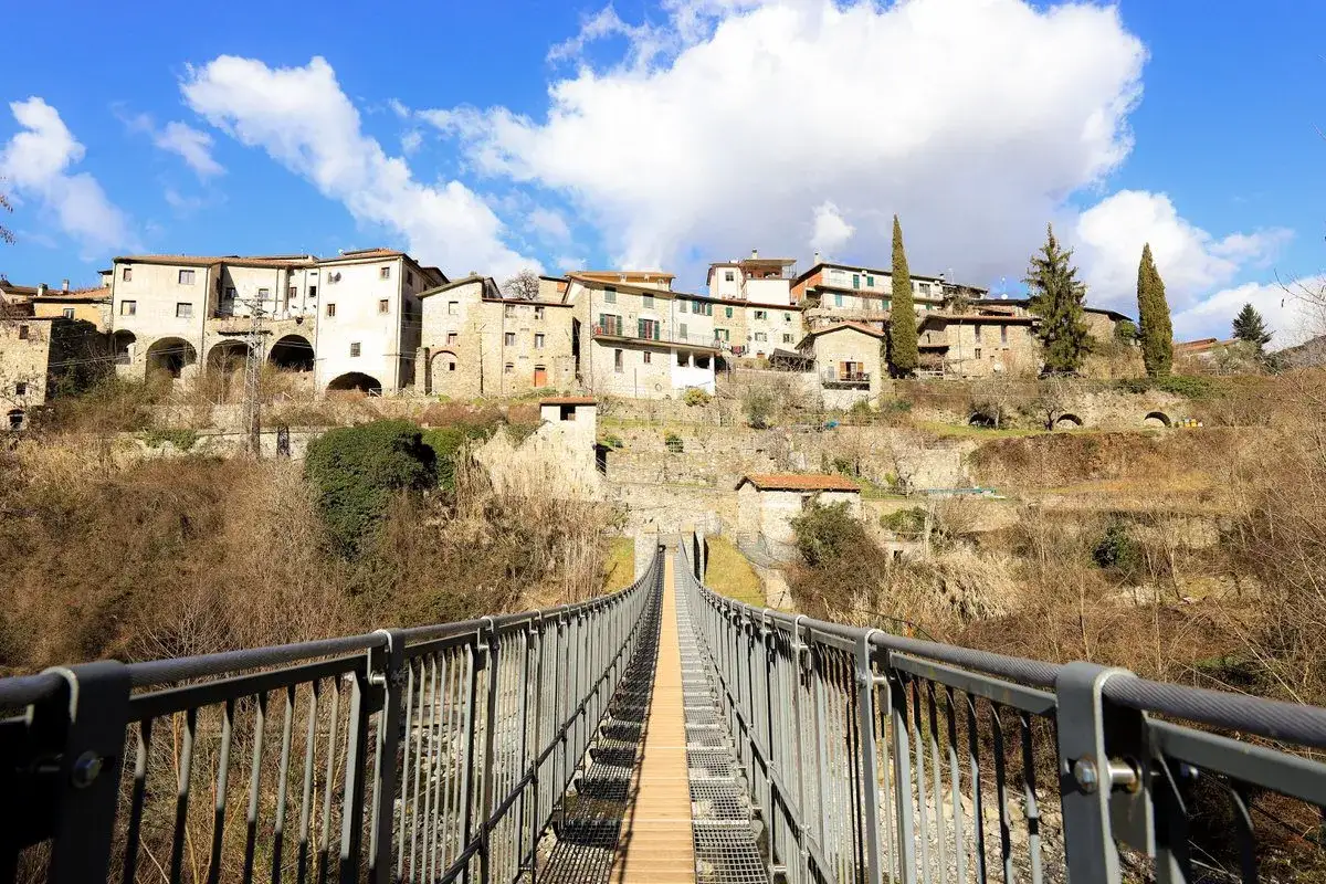 Ponte sospeso che conduce al borgo di Casola in Lunigiana, con case in pietra arroccate sulla collina sotto un cielo azzurro e nuvoloso.