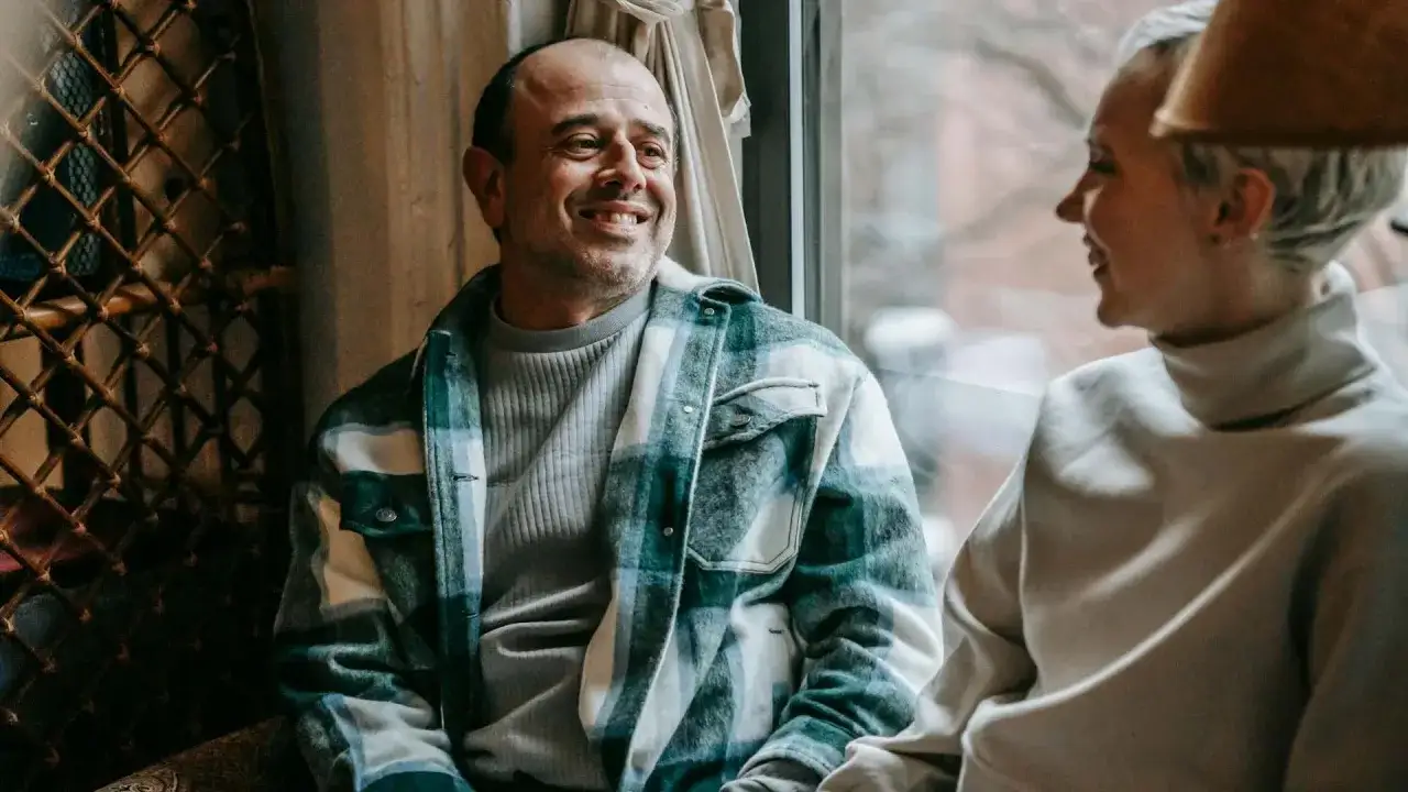 Coppia sorridente vicino a una finestra. Lui, con camicia a quadri, sembra raccontare qualcosa che suscita in lei una reazione di gelosia retroattiva.