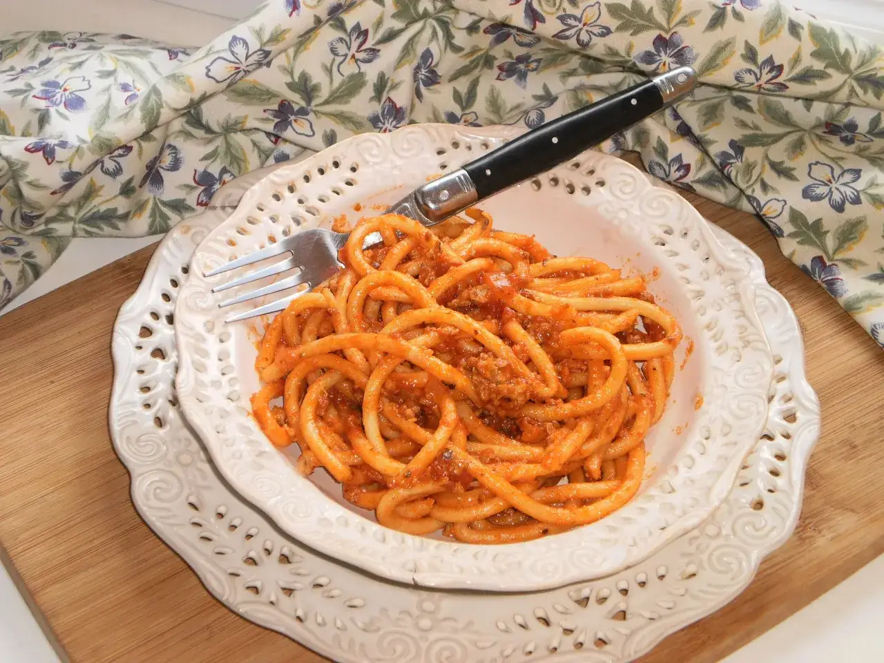 Talerz z makaronem bucatini w sosie bolońskim, obok widelec i serwetka w kwiaty. Czy to bucatini czy spaghetti?