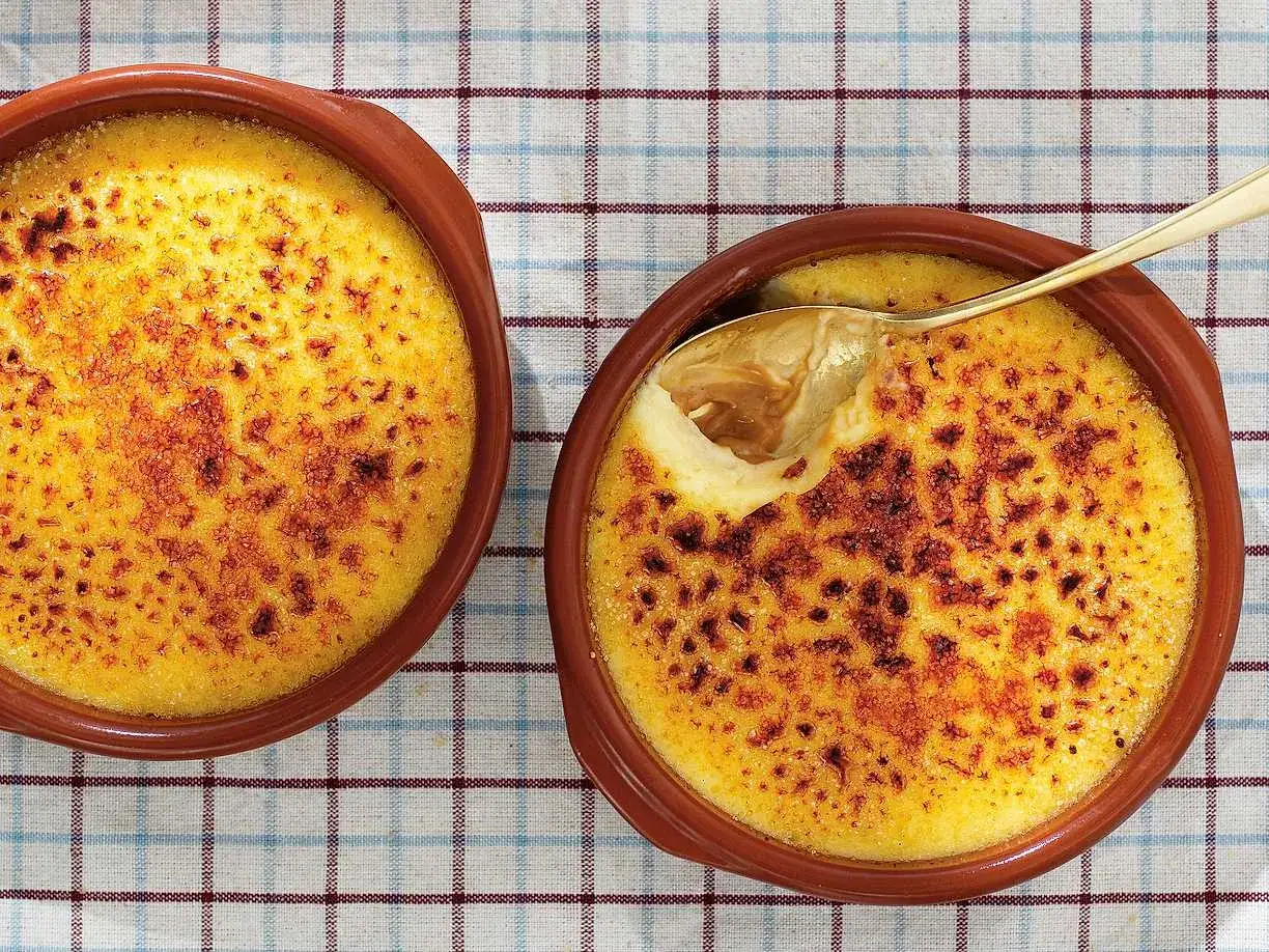 Tv&aring; kr&auml;miga creme brulee i terrakottaformar p&aring; en rutig duk. En sked har precis brutit igenom den karamelliserade ytan.