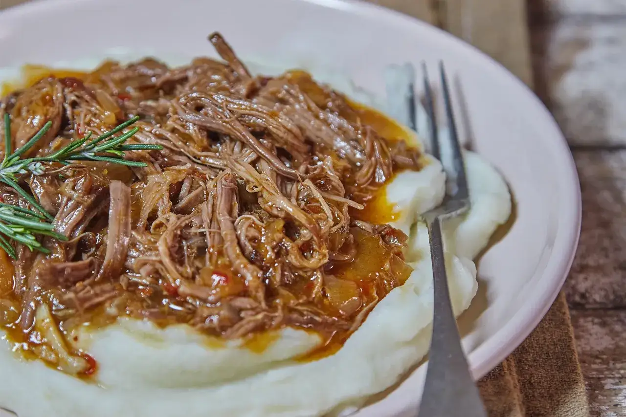 Carne mechada jugosa y deshebrada sobre puré de patatas cremoso, decorada con romero fresco. Un plato reconfortante.