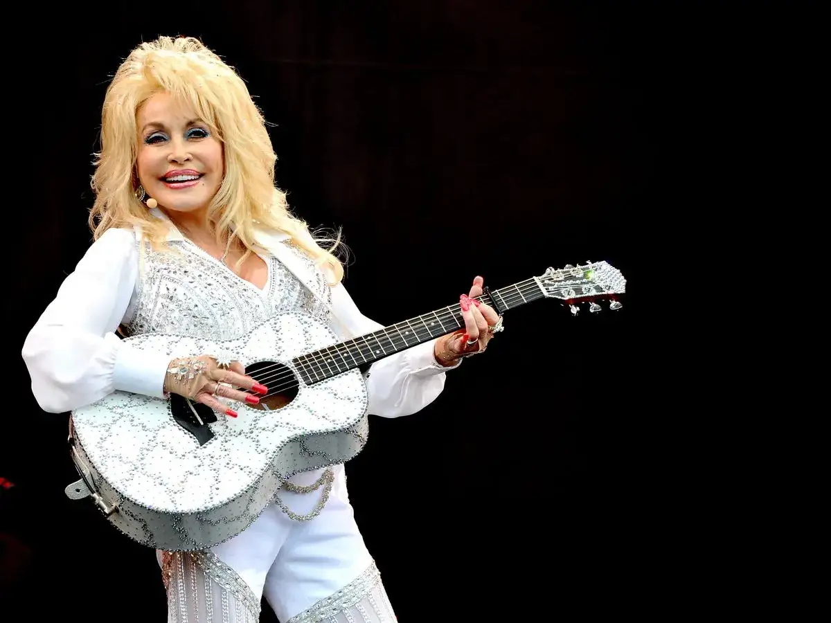 Dolly Parton, w białym, błyszczącym stroju, gra na gitarze. Wyobraź sobie, że śpiewa